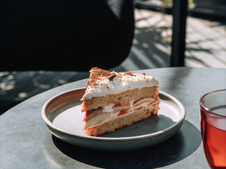 Ein Stück Apfelkuchen mit Haselnussboden, Apfelfüllung, Sahne und Kakaopulver auf einem Teller